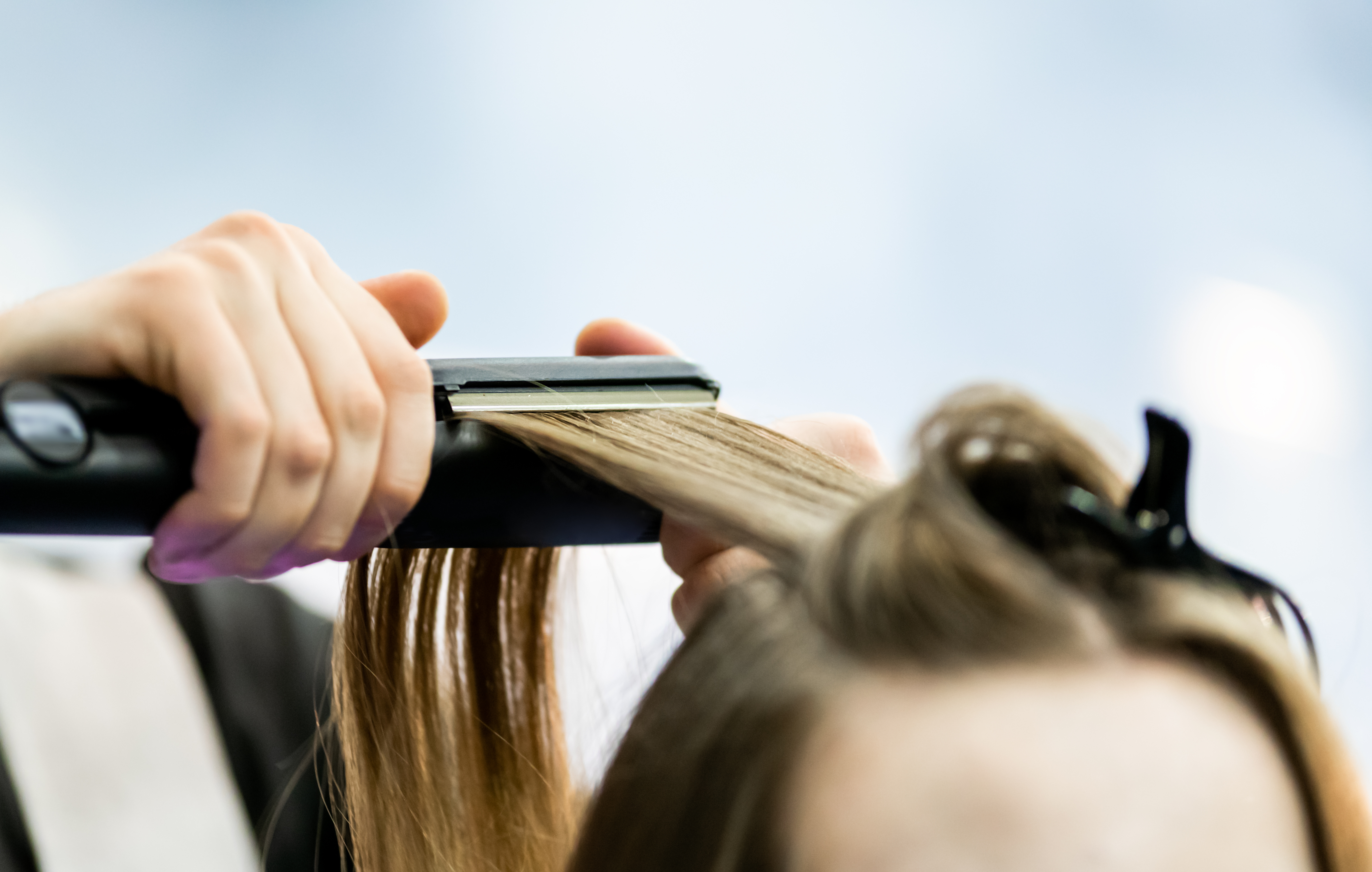 close-up of hands using a flat iron to straighten light brown hair section in a salon