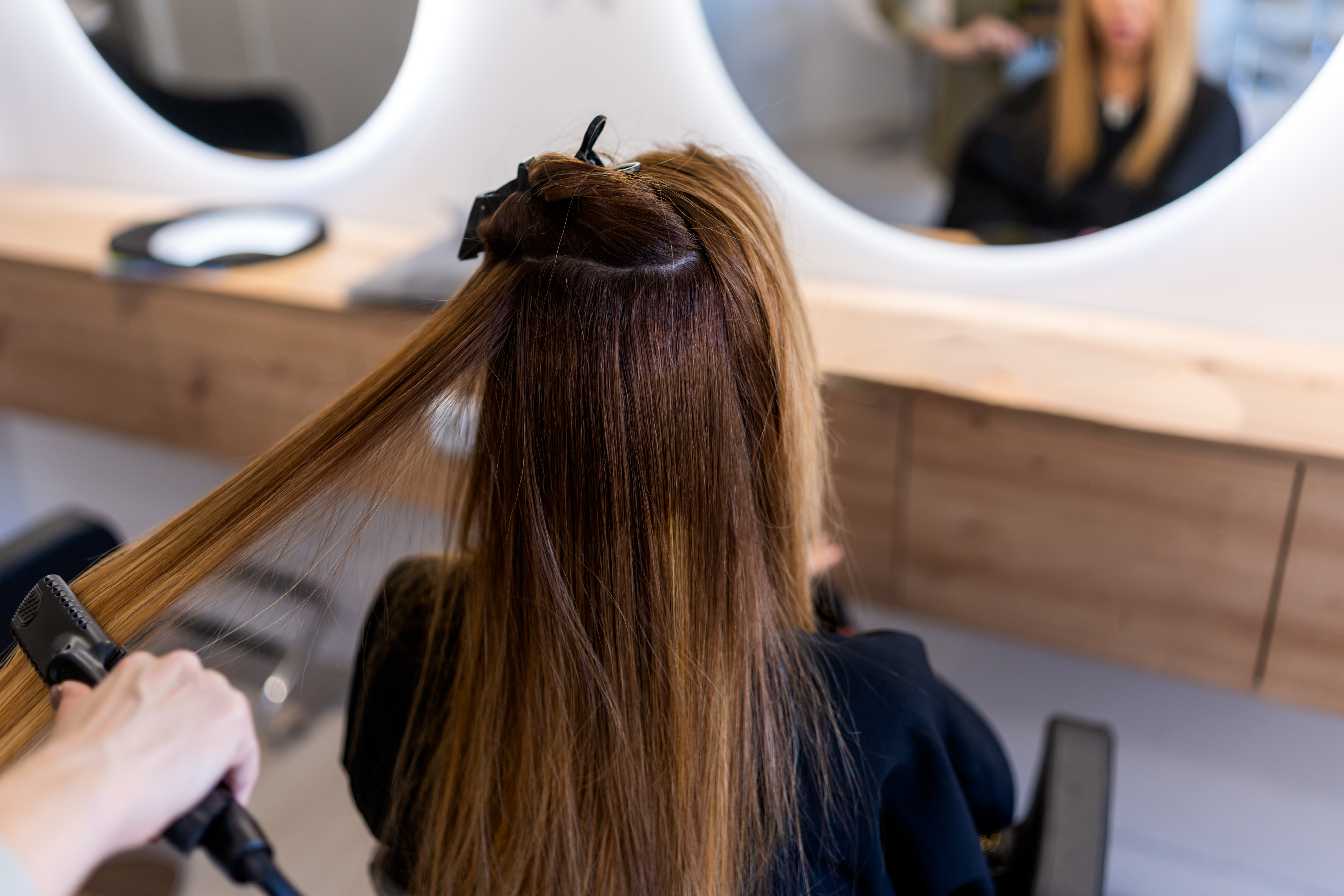 hairstylist using a flat iron on long brown hair in a salon with a mirror and wooden counter visible
